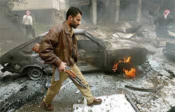 Un iraquí armado pasa delante de un coche ardiendo tras la explosión de dos bombas en un mercado de Bagdad.