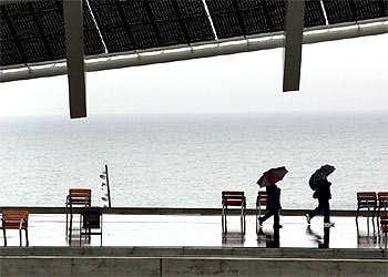 Dos visitantes pasan, paraguas en mano, bajo la placa fotovoltaica del recinto Fórum.