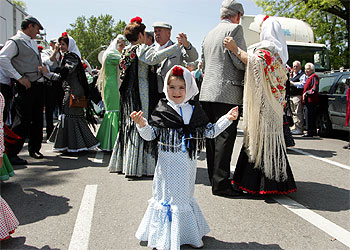 Una niña luce su vestido de chulapa en la pradera de San Isidro. A su lado, los mayores bailan un chotis.