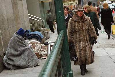 Una mujer con abrigo de piel pasa al lado de un mendigo arropado con mantas en la calle 51.