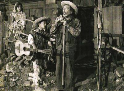  Ojitos  (Mario Ramírez) asiste a una actuación del ciego Don Carmelo (Miguel Inclán), en presencia de Meche (Alma Delia Fuentes), en  un fotograma de  Los olvidados. 