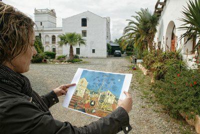 Una joven muestra una reproducción de  La masia,  de Miró, ante el escenario real de la pintura, que se convertirá en museo.